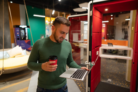 Bearded man holding laptop and drinking coffee in open space office.