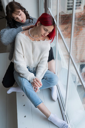 Two women dressed in sweaters sit by the window and gently hug. Lesbian intimacy.の写真素材