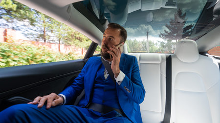 A caucasian man in a blue suit is talking on the phone in the back seat of a car. Business class...