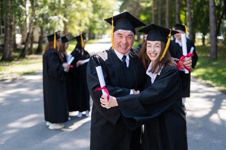 A group of graduates in robes outdoors. An elderly man and a young woman congratulate each other on their graduation.の写真素材