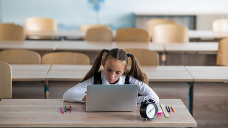 Caucasian girl sits at a desk at school and studies at a laptop. Alarm clock on the table.