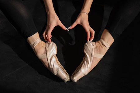 A faceless ballerina sits on the floor and makes a heart shape with her hands and pointe shoes. Top...