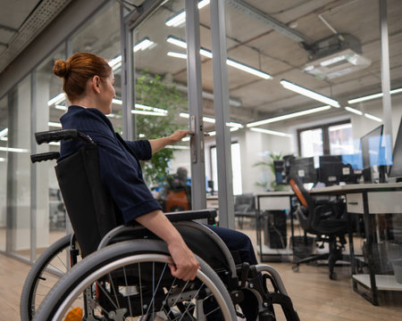 Red-haired caucasian woman in a wheelchair trying to open the door in the office.の写真素材