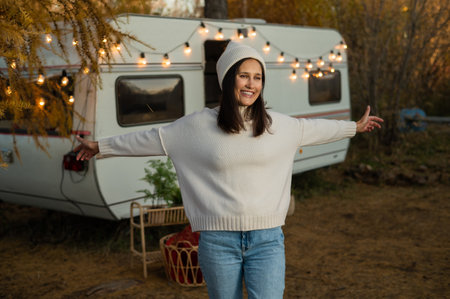 A pensive Caucasian woman in a white knitted sweater and a hat is resting near a motorhome in a warm autumn. Travel by camper.の写真素材
