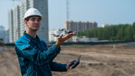 A man in a helmet and overalls controls a drone at a construction site. The builder carries out technical oversight.の写真素材