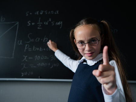Portrait of a caucasian girl in glasses in the classroom. The schoolgirl writes the formula with chalk on the blackboard and looks into the camera.の写真素材