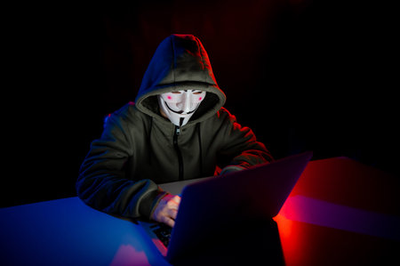 June 5, 2022 Novosibirsk, Russia: Anonymous in a hood is typing on a laptop in the dark in red blue...