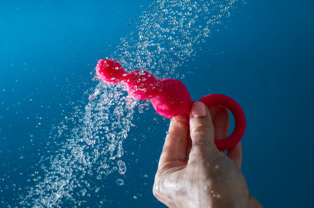 Woman holding pink anal beads under running water on blue background. Sex toy hygiene concept.の写真素材