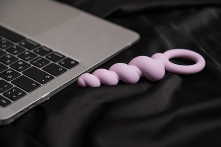 Close-up of lilac anal beads next to laptop on black silk sheet.の写真素材