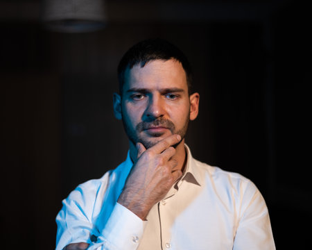 A bearded man stands in the dark and looks ahead puzzled.
