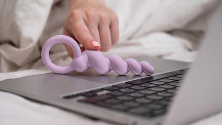 Woman holding lilac anal beads next to laptop while lying on bed.の写真素材