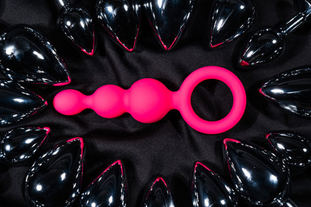 Pink anal beads among many metal butt plugs on a black silk sheet.の写真素材