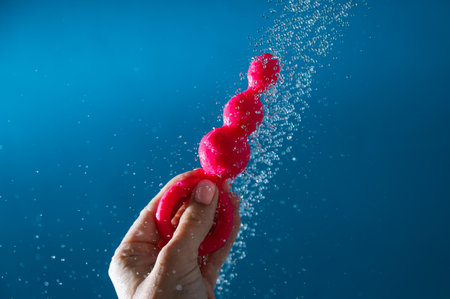Woman holding pink anal beads under running water on blue background. Sex toy hygiene concept.の写真素材
