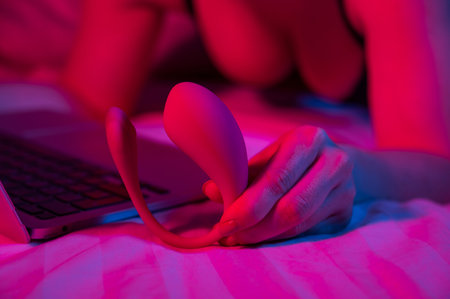 A woman is lying in bed holding a curved vibrator and looking at a laptop. Girl using sex toy in blue-red light.の写真素材