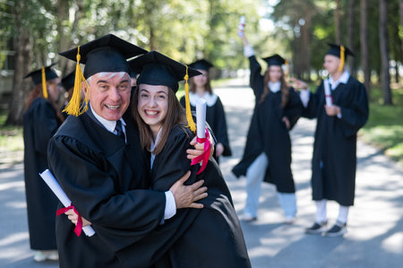 A group of graduates in robes outdoors. An elderly man and a young woman congratulate each other on their graduation.の写真素材