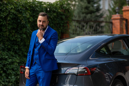 Caucasian bearded man in a blue suit stands near a black car in the countryside in summer.の写真素材