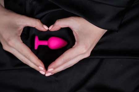 Woman making a heart shape with her palms around a pink butt plug on a black silk sheet.の写真素材