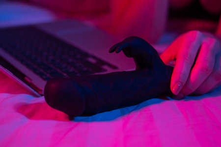 A woman lies in bed with a black dildo and looks at a laptop. Girl using sex toy in blue-red light.の写真素材