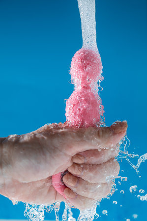 Woman holding pink anal beads under running water on blue background. Sex toy hygiene concept.の写真素材
