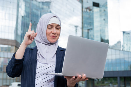 Business woman in hijab and suit is using a laptop and holding her index finger up outdoors.の写真素材