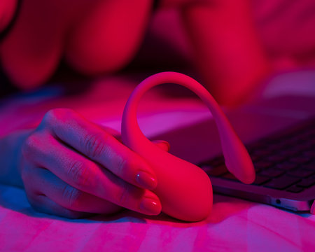 A woman is lying in bed holding a curved vibrator and looking at a laptop. Girl using sex toy in blue-red light.の写真素材