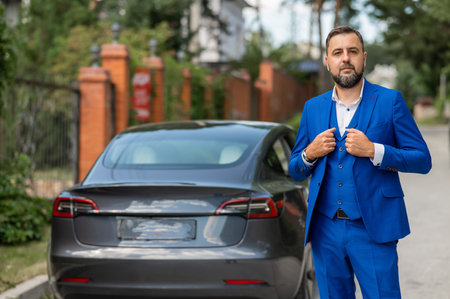 Caucasian bearded man in a blue suit stands near a black car in the countryside in summer.