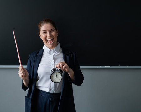 Caucasian female teacher in a pantsuit holds a pointer and a school alarm clock near the blackboard.の写真素材