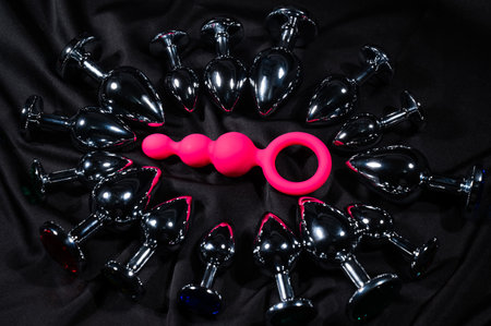 Pink anal beads among many metal butt plugs on a black silk sheet.の写真素材