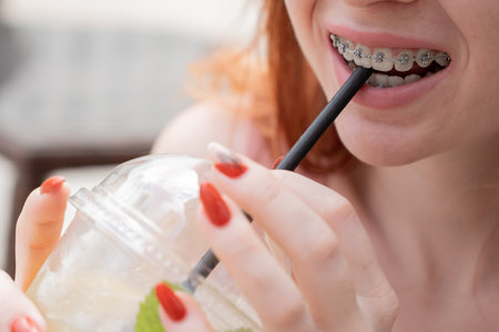 Young beautiful red-haired woman with braces drinks cooling lemonade outdoors in summer. Portrait of...