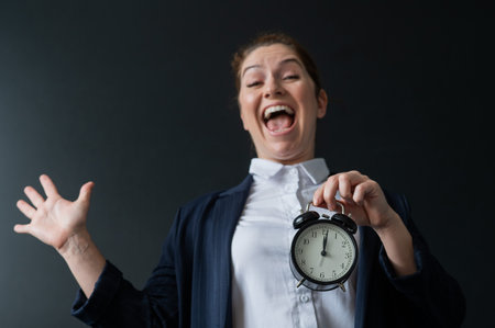 A happy female teacher in a pantsuit holds an alarm clock standing at the blackboard and rejoices at the end of the lessons at school.の写真素材