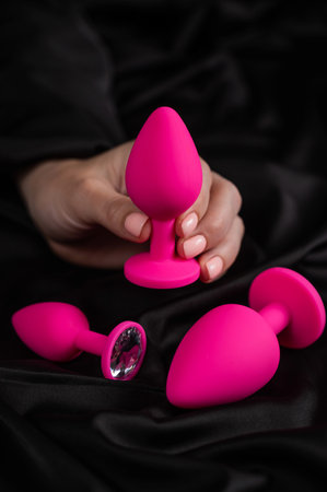 A woman is holding a pink butt plug on a black silk sheet.