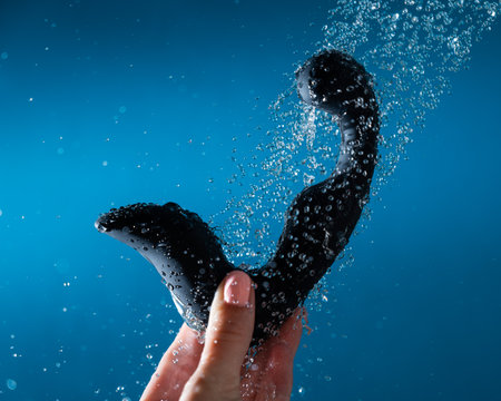A woman washes a black prostate stimulator under running water on a blue background. Sex toy hygiene concept.の写真素材