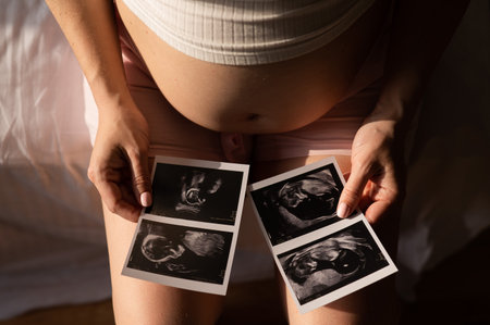 A pregnant woman looks at a photo from an ultrasound while sitting on the bed. Close-up of a pregnant womans belly.の写真素材