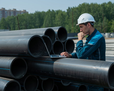 Caucasian male builder in a hard hat stands near the pipes and uses a laptop at a construction site.の写真素材