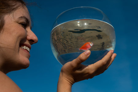 Woman holding round aquarium with goldfish on blue sky background.の写真素材