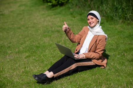 A young woman wearing a hijab sits on a lawn and uses a laptop outdoors and shows a thumbs up.の写真素材