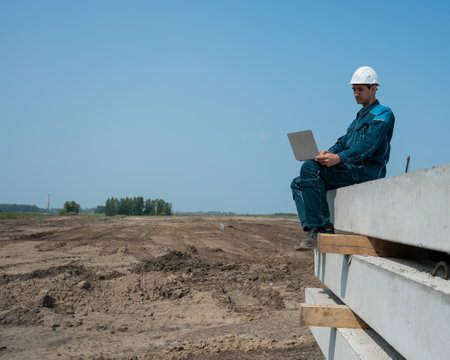 Caucasian male builder in hardhat sits on floor slabs and uses laptop at construction site.の写真素材