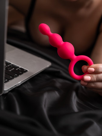 Woman holding pink anal beads next to laptop while lying on black sheet.の写真素材