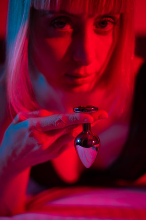 The blonde lies on the bed in red blue neon light and holds a steel butt plug.の写真素材
