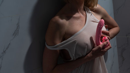 A woman in a white tank top stands in the shower and holds a curved pink sex toy.の写真素材