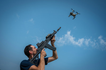 A man aims to shoot a rifle at a flying drone against a blue sky.の写真素材