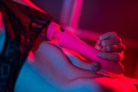 A faceless woman lies in a bedroom in a blue-red neon light and uses a pink vibrator. Close-up of the abdomen.の写真素材