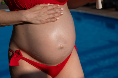 Close-up of the belly of a pregnant woman sunbathing in a red bikini. Expectant mother applies sunscreen.の写真素材