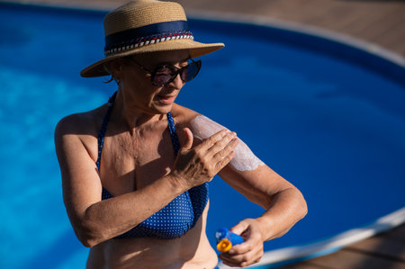 Portrait of an old woman in a straw hat, sunglasses and a swimsuit applying sunscreen to her skin while relaxing by the pool.の写真素材