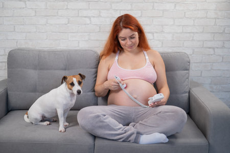 A red-haired pregnant woman sits on a sofa with a jack russell terrier dog and uses a home doppler.の写真素材