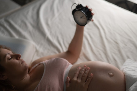A pregnant woman lies in bed and holds an alarm clock.