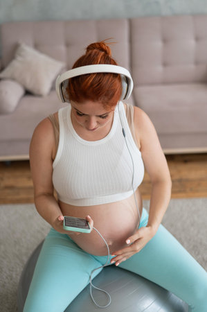 Caucasian red-haired pregnant woman sitting on fitball and listening to music with headphones at home.の写真素材