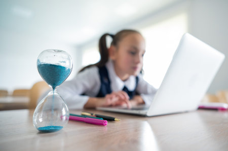 Caucasian girl sits at a desk at school and studies at a laptop. Hourglass on the table.の写真素材