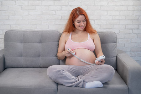 Red-haired pregnant woman sits on the couch and uses a home dopplerの写真素材