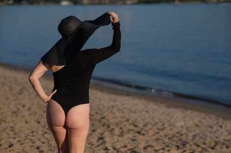 Rear view of woman in large straw hat and black swimsuit posing on the beach